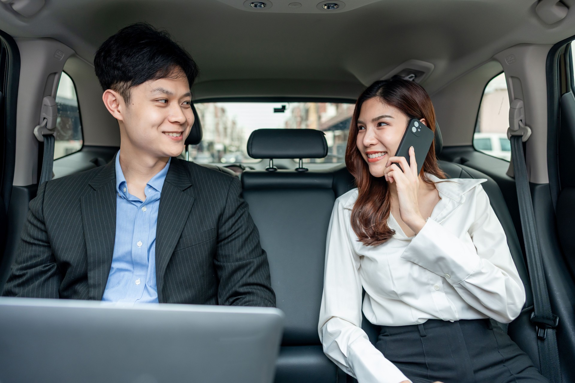 Asian businessman and woman working while sitting in back seat of a car. Attractive young colleagues work remotely or checking information on device, enjoy productive business commute through the city